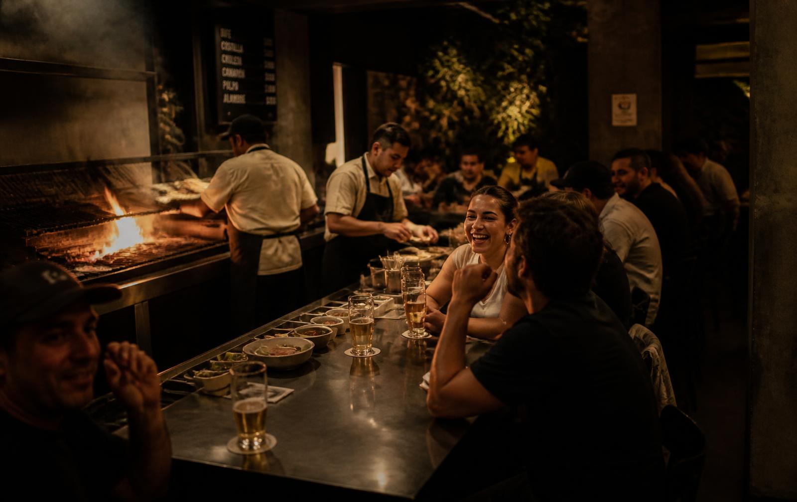 Gente riendo y tomando cerveza en la barra de Los Popis frente a la parrilla de carbón abierta